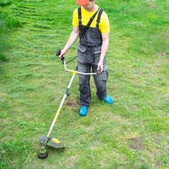 A male gardener mows the green grass of the lawn in the backyard with a gasoline mower. Trimmer for the care of a garden plot