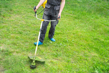 A male gardener mows the green grass of the lawn in the backyard with a gasoline mower. Trimmer for the care of a garden plot