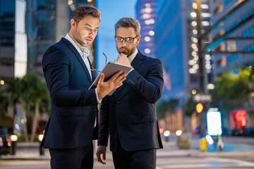 Business man in night Ney York. Business people discuss a project. The two business teams men team using tablet laptop outdoor. Businessmen looking tablet with business success in night city.