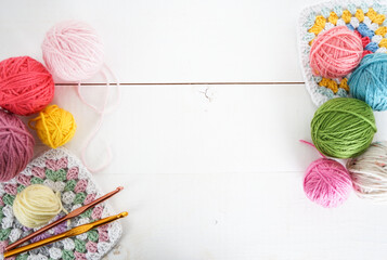multicolored woolen balls with granny sqaure and crochet hook on white wooden ground with space for...