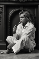Pensive middle aged 45 years old woman sitting on floor in dark living room, lighting cigarette. Thoughtful sad alone mature lady in white, indoors. Loneliness emotion concept. Copy text space for ad
