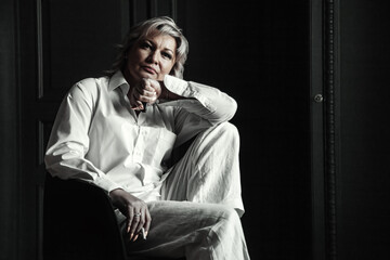 Black white portrait of pensive middle aged 45 years old woman with cigarette sitting on chair in dark room, looking at camera. Thoughtful sad alone mature lady. Aging concept. Copy ad text space