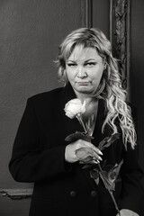 Black white portrait of pensive middle aged 45 years old stylish woman with white rose posing, looking at camera. Thoughtful sad alone mature lady standing at black wall. Emotion concept. Copy space