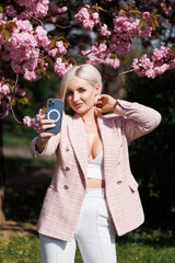 A beautiful blonde is posing straight with a smartphone taking pictures under the sun's rays on a beige background and a branch of blooming sakura.