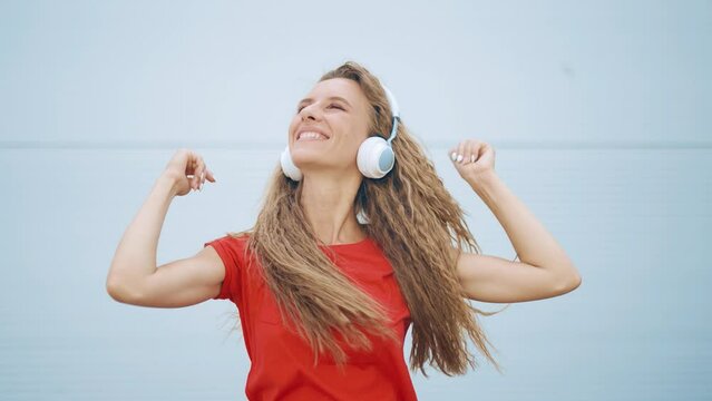 A young girl in headphones is listening to music and amusingly dancing on a light background. Woman is having fun alone and feeling happy and carefree. Cheerful dance. Entertainment and joy concept.