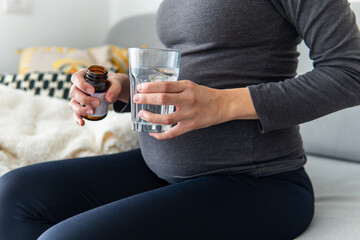 Pregnant woman taking vitamins pills and minerals from a jar with a glass of water while sitting comfortably on the sofa. Pregnancy, healthcare, and supplements concept. Woman taking medicine at home.