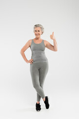 full length of cheerful and retired woman in sportswear posing with hand on hip and showing like on grey.