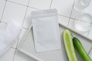Facial sheet mask packaging mockup with fresh cucumber, towel and glass cup on white tile...