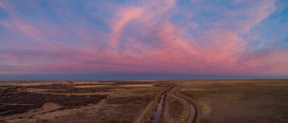 Aerial panoramic shot of a field with a narrow lake on it during a pink sunset