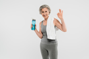 happy senior woman with white towel on shoulder holding sports bottle with water and showing ok isolated on grey.