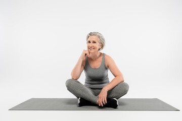 full length of pleased senior woman in wireless earphones listening music and sitting in yoga pose on grey.