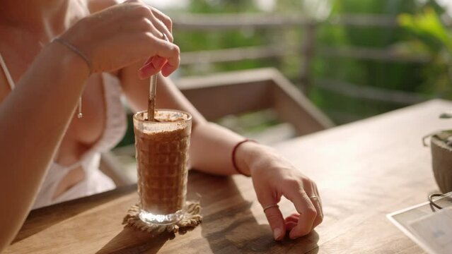 Female hands stirring cocktail with metal straw in outdoor restaurant. Young woman drinking chocolate milkshake in sunny tropical cafe. Girl at the table having iced coffee in a tall glass
