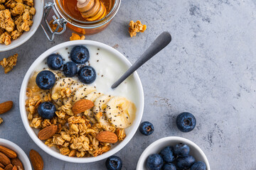 Bowl of greek yogurt with oatmeal granola, banana, blueberry, nuts, chia seeds and honey on gray table. Top view