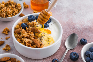 Pouring honey into the bowl of greek yogurt with oatmeal granola, banana, blueberry, nuts, chia seeds.