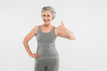 cheerful and retired woman in sportswear posing with hand on hip and showing like isolated on grey.