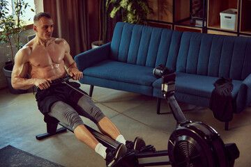 Muscular young man training shirtless with stationary rowing machine, doing exercises at home on...