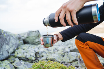 Pour the tea from the thermos into the mug with your hand, the water flows hot coffee into the glass. A tourist drinks coffee in the mountains, camping in nature, hands with a cup, food on a hike.
