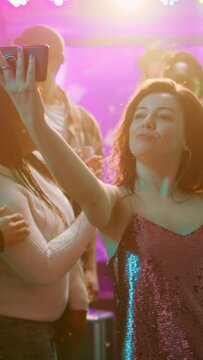 Vertical video: Happy girl taking pictures on phone, recording video for memories to have fun at disco party event. Woman partying with group of friends and taking photos, filming on smartphone
