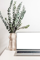 Laptop mockup on the table with vase and green eucalyptus leaves. Laptop with white screen for study, home office, website promotion, business branding, aesthetic style