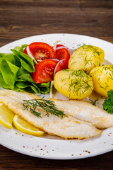 Fish dish - fried cod with boiled potatoes and fresh vegetables on wooden table
