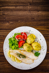Fish dish - fried cod with boiled potatoes and fresh vegetables on wooden table
