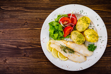 Fish dish - fried cod with boiled potatoes and fresh vegetables on wooden table
