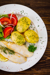 Fish dish - fried cod with boiled potatoes and fresh vegetables on wooden table
