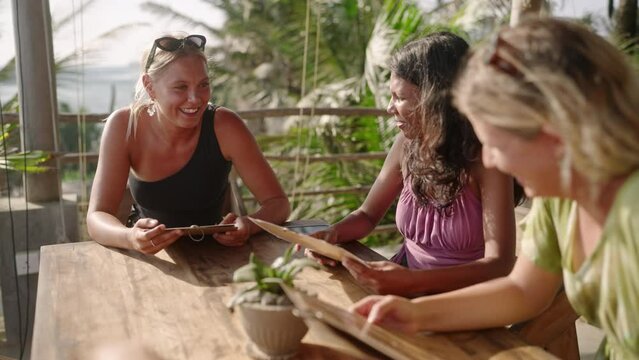 Diverse female friends in outdoor restaurant reading menu. Multiracial girls sitting at the table in a tropical cafe talking to each other choosing food. Young multiethnic women having dinner