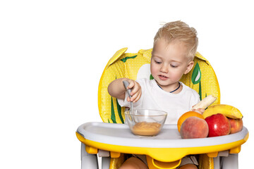 A cheerful and beautiful baby sits in a child seat and eats fruit puree on his own by a boat in the kitchen