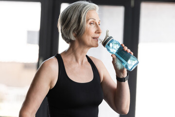 senior woman with grey hair drinking water from sports bottle in gym.