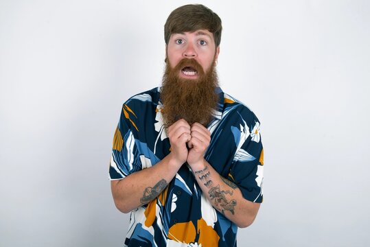Dreamy charming red haired man wearing printed shirt over white studio background with pleasant expression, keeps hands crossed near face, excited about something pleasant.