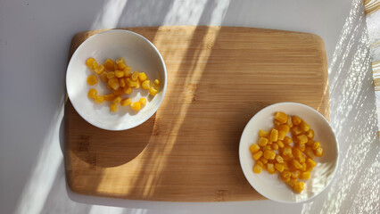 Lot of pieces of canned yellow corn on plate which is on wooden bamboo cutting board on white background. The concept of cooking and delicious healthy food