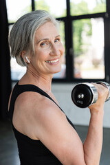 Fototapeta na wymiar overjoyed senior woman in wireless earphone working out with dumbbell in gym.