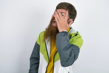 red haired man wearing sportswear standing over white studio background makes face palm and smiles broadly, giggles positively hears funny joke poses