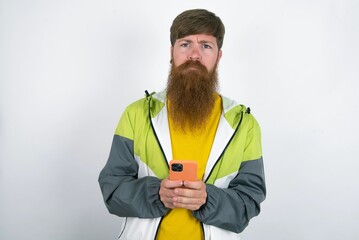 Upset dissatisfied red haired man wearing sportswear standing over white studio background uses mobile software application and surfs information in internet, holds modern mobile hand