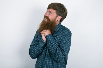 Happy red haired man wearing plaid shirt over white studio background anticipates something awesome happen, looks happily aside, keeps hands together near face, has glad expression.