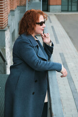 Attractive red-haired woman in a blue coat, jeans and a loose shirt stands outside a brick building with mirrored door. A woman in sunglasses looks away. Vertical portrait of woman