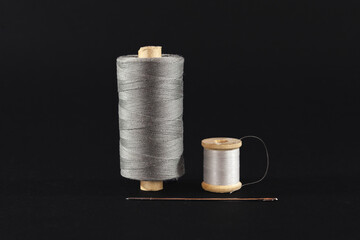 Studio shot of a grey and white thread reel, one big one small size, & a sewing needle isolated on black background. Spool (or reel), thread & needle are used for DIY textile sewing & fashion creation