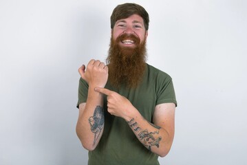 red haired man wearing green T-shirt over white studio background In hurry pointing to wrist watch, impatience, looking at the camera with relaxed expression