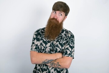 Dissatisfied red haired man wearing printed shirt over white studio background purses lips and has unhappy expression looks away stands offended. Depressed frustrated model.