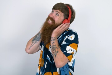 Joyful red haired man wearing printed shirt over white studio background sings song keeps hand near mouth as if microphone listens favorite playlist via headphones