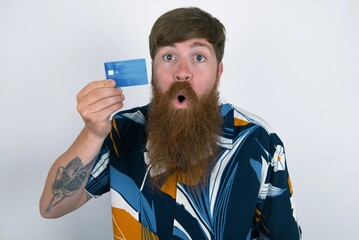 Photo of red haired man wearing printed shirt over white studio background amazed shocked hold credit card payment