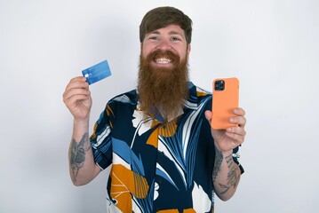 red haired man wearing printed shirt over white studio background wears stereo headphones on ears holds modern mobile phone and credit card