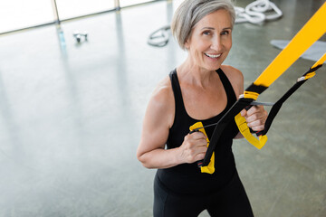 high angle view of happy senior woman in black sportswear working out with suspension straps in gym.