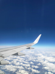 View from the window of the wing of an airplane flying high above a blue sky. You can see the coluds and cities below us.