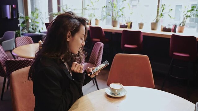 Female blogger making photo of cup coffee for social networks while sitting in cozy coffee shop. Smiling pretty lady using phone for content. Social networks concept. Real time