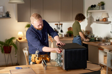 Mature handyman using his work tools to repair oven at home with housewife doing housework in...