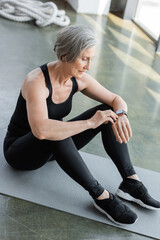 Fototapeta na wymiar full length of fit senior woman in black leggings and tank top checking fitness tracker while sitting on fitness mat.