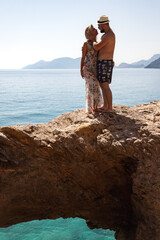 Lovely couple woman and man standing hugging on rock at azure sea background, summer vacation. Happy young family enjoying honeymoon on seacoast together. Travel holidays concept. Copy ad text space