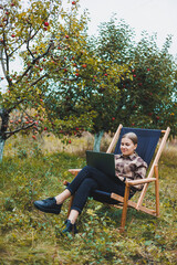 A cute young female freelancer is working in the garden outdoors while sitting in a comfortable garden chair. Remote work on a portable laptop